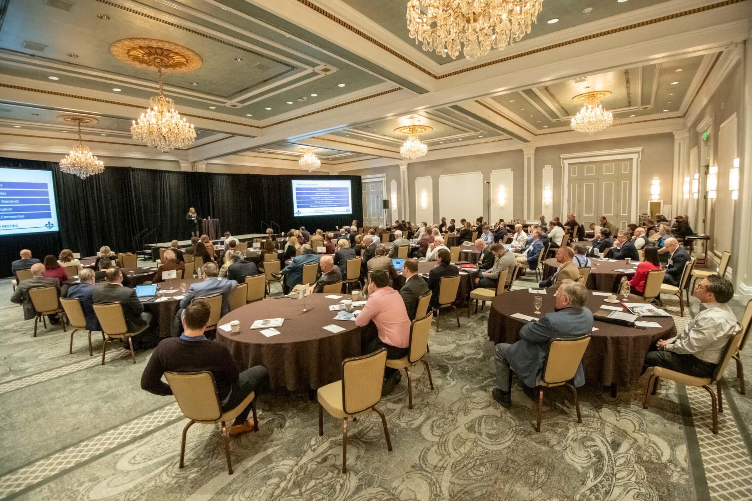 A woman on stage addresses a crowded ballroom of meeting attendees at round tables